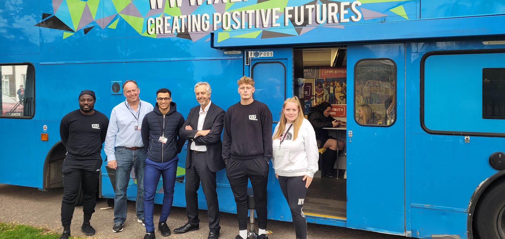 A group of six stand outside XLP's blue double-decker youth bus while young people sit inside at a table
